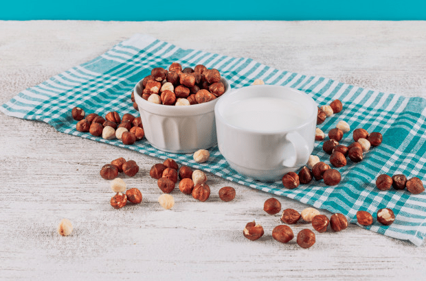 Picture-of-a-hazelnut-milk-cup-and-hazelnuts-spread-across-a-blue-cloth-with-a-white-background
